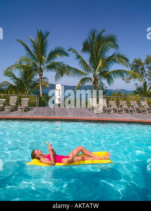 Woman relaxing on a yellow pool float in a swimming pool with palm trees and ocean in the background Stock Photo