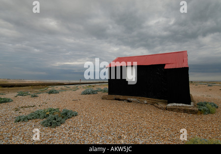 Red roofed and black corrugated iron hut on the shingle above the mouth of the River Rother Stock Photo