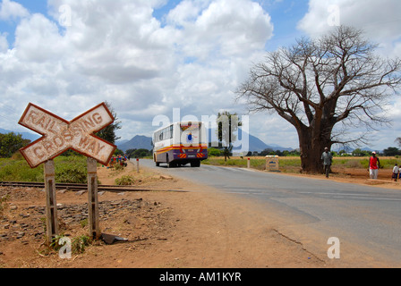 Sign Railway Crossing with long distance bus on country road and baobab (Adansonia digitata) near Moshi Tanzania Stock Photo
