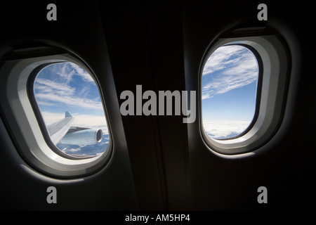 View from plane. Two windows like eyes looking out over wing and jet engine. White clouds underneath; contrails in blue sky. Stock Photo