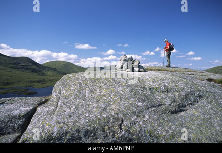Hill walking in the Galloway Hills hill walker on huge stone of granite by cairn on Craig Neldricken with Loch Enoch behind UK Stock Photo