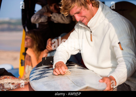 Man waxing surfboard with couple smiling in background. Stock Photo