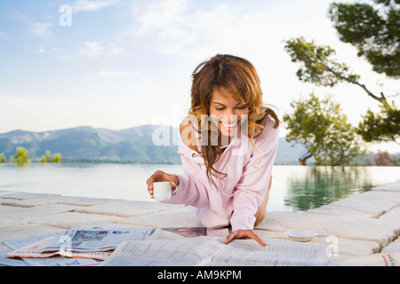 Woman smiling by infinity pool reading newspaper and drinking coffee. Stock Photo