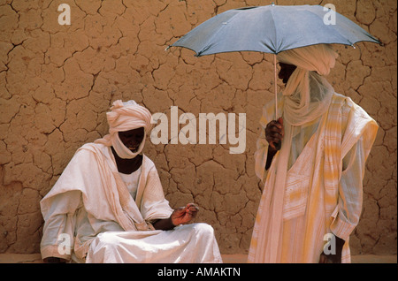 Two men in traditional white robes and turbans chatting outside an adobe mud wall in a village in Mali Africa Stock Photo