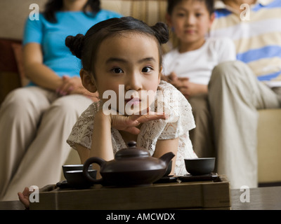 Girl in living room with tea pot and cups with parents and brother in background Stock Photo