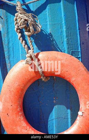Orange lifebuoy swinging on a frayed rope attached to the helm of a blue boat in bright sunshine. Stock Photo