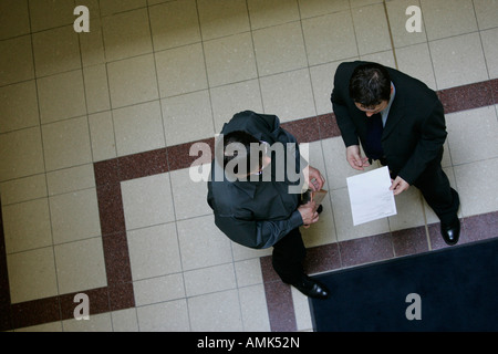 TWO MALE OFFICE WORKERS HAVING A DISCUSSION IN A TILED CORRIDOR SEEN FROM ABOVE. Stock Photo