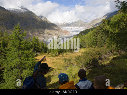 A guide points out features to visiting school children of the Great Aletsch Glacier beyond valley with trees Valais Switzerland Stock Photo