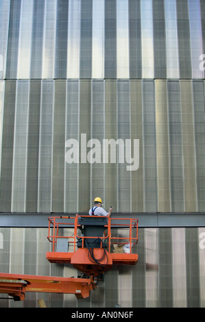 Construction worker wearing yellow hat on orange elevated crane platform in front of metal surface of a skyscraper New York USA Stock Photo
