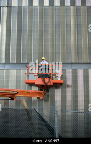 Construction worker wearing a yellow hat stands on an orange elevated crane platform in front of the metal surface of skyscraper Stock Photo