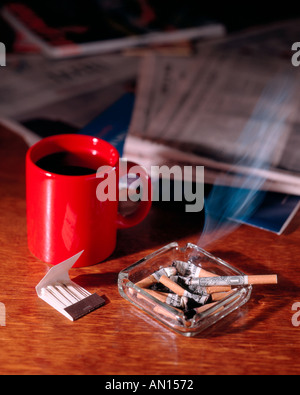 Burning money wrapped cigarette going up in smoke in overflowing ashtray with book of matches and newspaper and red coffee mug Stock Photo