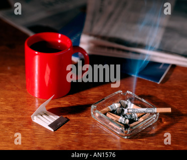 Burning money wrapped cigarette going up in smoke in overflowing ashtray with book of matches and newspaper and red coffee mug Stock Photo