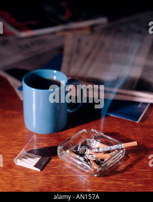 Burning money wrapped cigarette going up in smoke in overflowing ashtray with book of matches and newspaper and blue coffee mug Stock Photo