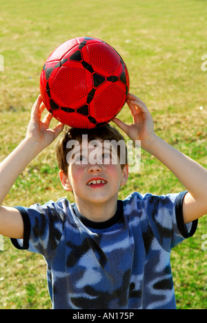 Young cute happy boy balancing a red soccer boy on his head outside Stock Photo