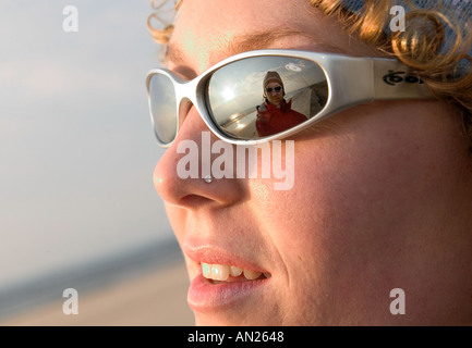An attractive young lady with a nose piercing dons stylish sunglasses, capturing the reflection of a man in a red jacket Stock Photo
