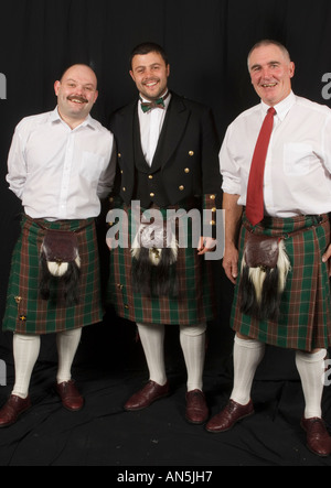 Three smiling men wearing  welsh tartan  kilts in front of black backdrop, Wales UK Stock Photo