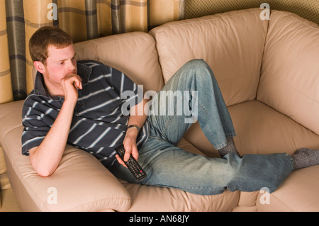 Man in his twenties holding a TV remote control whilst relaxing on a sofa Stock Photo