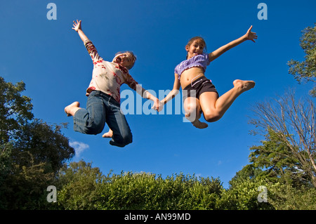 Horizontal portrait of two young caucasian girls in mid air against a blue sky, bouncing on a trampoline. Stock Photo