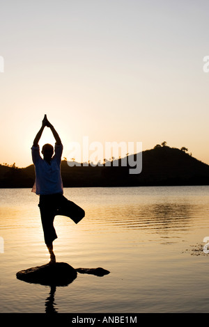 Silhouette of a man in a Hatha Yoga posture on a rock in a lake at sunset Stock Photo