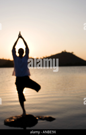 Silhouette of a man in a Hatha Yoga posture on a rock in a lake at sunset Stock Photo