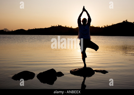 Silhouette of a man in a Hatha Yoga posture on a rock in a lake at sunset Stock Photo