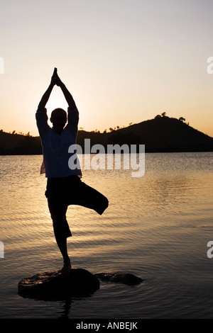 Silhouette of a man in a Hatha Yoga posture on a rock in a lake at sunset Stock Photo