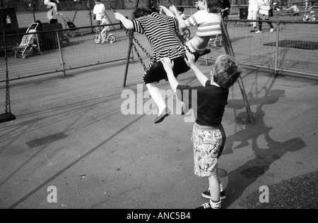 Black and white of a small boy pushing his sister on a swing in a children's playground Stock Photo