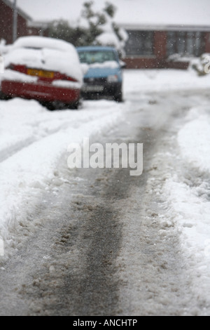 tyre tracks and car marks in the middle of the road of snow covered street with ice slush and parked cars Stock Photo