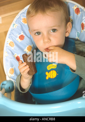 Young boy sitting down in a highchair eating a chocolate roll Stock Photo