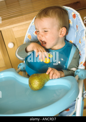 Young boy sitting down in a highchair refusing to eat a pear Stock Photo