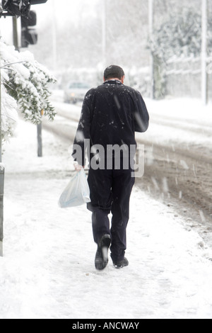 man with no hood walking uphill along snow covered footpath carrying plastic bag looking down at phone Stock Photo
