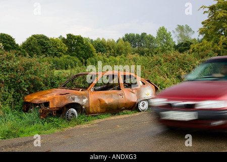 Car passes a burnt out wreck by the roadside Oxfordshire United Kingdom Stock Photo