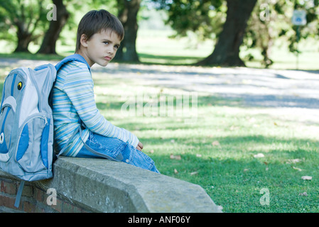 Boy sitting on low wall, wearing backpack Stock Photo