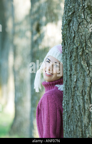 Young woman wearing knit hat, leaning against tree, smiling over shoulder at camera Stock Photo