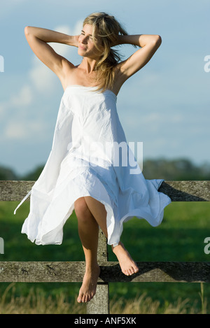 Young woman sitting on top of wooden fence, hands behind neck, smiling Stock Photo