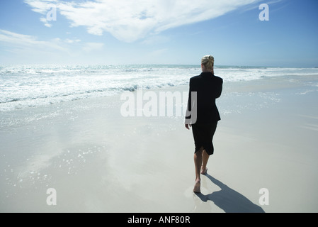 Businesswoman using cell phone, walking barefoot across beach, full length, rear view Stock Photo