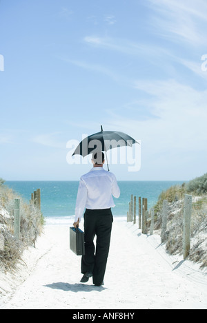 Businessman walking on sandy path leading to ocean, using umbrella, rear view Stock Photo