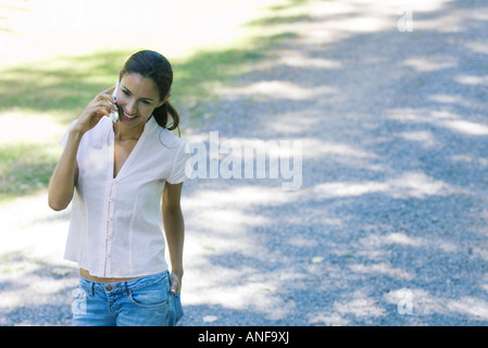 Casually dressed woman using cell phone, smiling as she walks through park Stock Photo