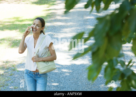 Casually dressed woman using cell phone, smiling as she walks through park, purse on shoulder Stock Photo