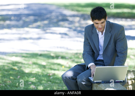 Businessman sitting on low wall in park, using laptop Stock Photo