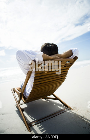 Man sitting in chair on beach hands, hands behind head, rear view Stock Photo