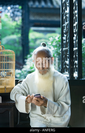 Elderly man wearing traditional Chinese clothing, using cell phone, next to bird cage Stock Photo