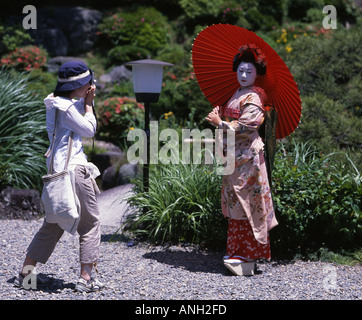 Tourist taking photo of woman dressed as maiko, apprentice geisha, holding red parasol in Japanese garden, Gion, Kyoto, Japan Stock Photo