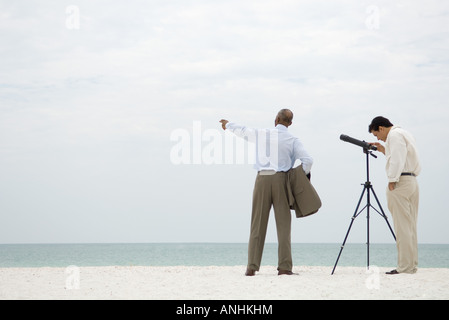 Two businessmen standing at the beach, one looking through telescope, the other pointing Stock Photo