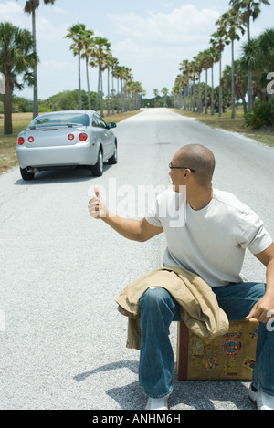 Male hitchhiker sitting on suitcase on road, looking over shoulder at passing car Stock Photo