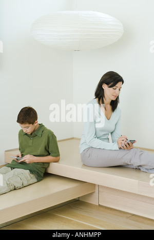 Woman and son sitting near each other, woman using PDA, boy playing with handheld video game Stock Photo