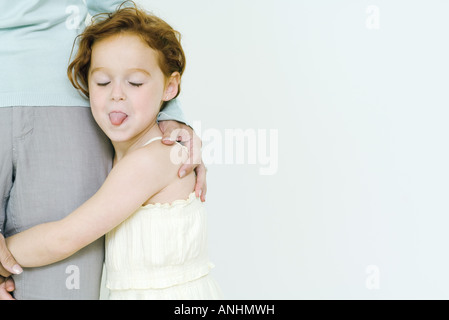 Little girl holding on to mother's leg, sticking tongue out at camera Stock Photo