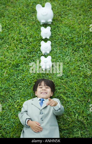 Boy dressed in suit lying on grass, laughing and pointing at camera, piggy banks above head Stock Photo