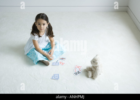 Girl sitting on the ground playing card game with teddy bear, looking up at camera Stock Photo