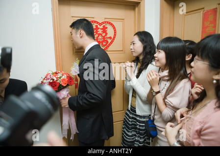 Chinese wedding, groom holding bouquet and knocking on bride's door while bride's friends watch Stock Photo